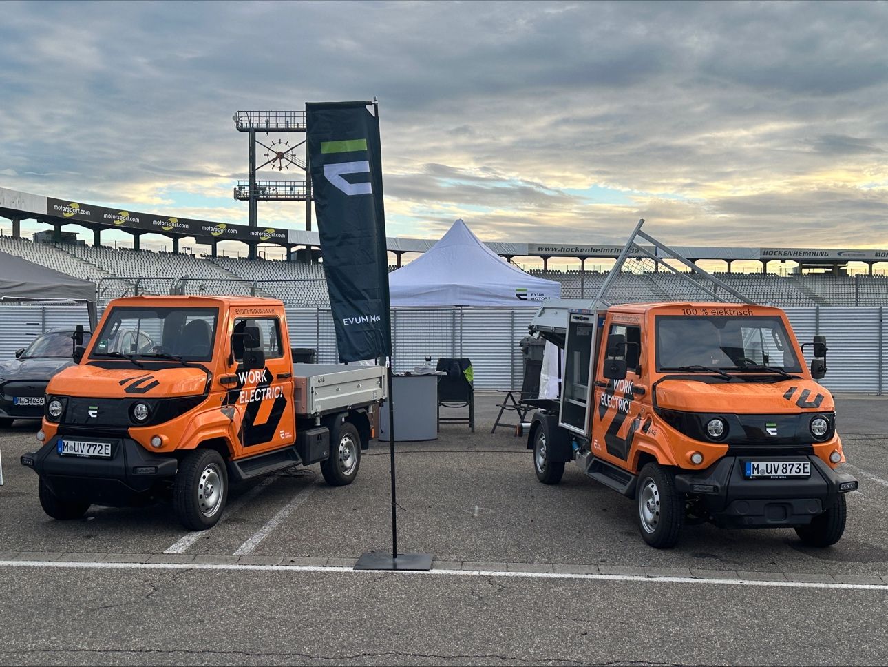 Zwei Evum-Nutzfahrzeuge stehen neben einer EVUM Motors-Flagge am Hockenheimring. Die Fahrzeuge symbolisieren die Förderung emissionsfreier Mobilität durch staatliche Förderprogramme. Foto: EVUM Motors.